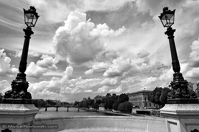 Looking from the Pont Neuf between two lanterns along the Seine, onto the Louvre and the Pont des Arts in front of spectacular clouds in the sky.